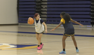 Two basketball players scrimmage on an indoor court. One player in a white practice jersey dribbles the ball while being guarded closely by another player in a dark practice jersey with yellow sleeves. The gym’s bleachers are visible in the background.