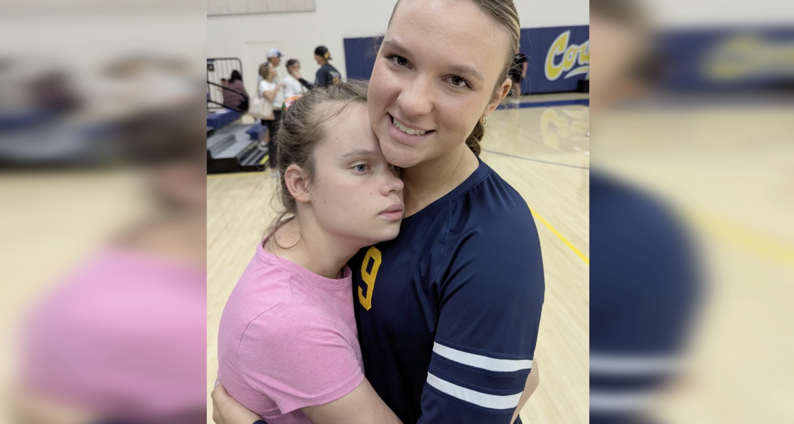 Two girls stand on a gymnasium floor, embracing closely. One girl, wearing a pink shirt, rests her head against the chest of an older girl in a navy sports jersey with the number 9, who is smiling gently at the camera. The background shows blurred gym equipment and a few people talking near the bleachers.