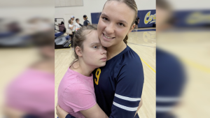 Two girls stand on a gymnasium floor, embracing closely. One girl, wearing a pink shirt, rests her head against the chest of an older girl in a navy sports jersey with the number 9, who is smiling gently at the camera. The background shows blurred gym equipment and a few people talking near the bleachers.