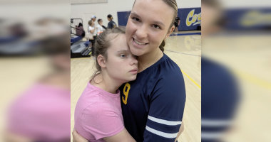 Two girls stand on a gymnasium floor, embracing closely. One girl, wearing a pink shirt, rests her head against the chest of an older girl in a navy sports jersey with the number 9, who is smiling gently at the camera. The background shows blurred gym equipment and a few people talking near the bleachers.