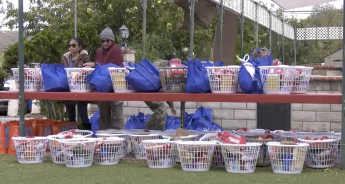 A row of white plastic baskets and blue reusable bags filled with food and household items are arranged on red shelves outdoors. Two people stand behind the top shelf, organizing the supplies. More filled baskets sit on the ground below, with trees and a brick wall in the backgr
