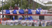 A row of white plastic baskets and blue reusable bags filled with food and household items are arranged on red shelves outdoors. Two people stand behind the top shelf, organizing the supplies. More filled baskets sit on the ground below, with trees and a brick wall in the backgr