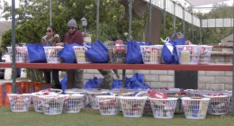 A row of white plastic baskets and blue reusable bags filled with food and household items are arranged on red shelves outdoors. Two people stand behind the top shelf, organizing the supplies. More filled baskets sit on the ground below, with trees and a brick wall in the backgr