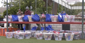 A row of white plastic baskets and blue reusable bags filled with food and household items are arranged on red shelves outdoors. Two people stand behind the top shelf, organizing the supplies. More filled baskets sit on the ground below, with trees and a brick wall in the backgr
