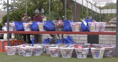A row of white plastic baskets and blue reusable bags filled with food and household items are arranged on red shelves outdoors. Two people stand behind the top shelf, organizing the supplies. More filled baskets sit on the ground below, with trees and a brick wall in the backgr