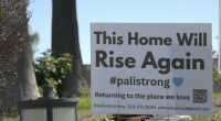 A white yard sign stands on a patch of grass in front of trees. The sign reads, “This Home Will Rise Again #palistrong” with a small blue heart. Below it says, “Returning to the place we love,” along with contact information and a QR code. A small outdoor light fixture sits in the foreground.