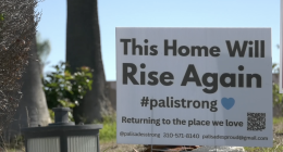 A white yard sign stands on a patch of grass in front of trees. The sign reads, “This Home Will Rise Again #palistrong” with a small blue heart. Below it says, “Returning to the place we love,” along with contact information and a QR code. A small outdoor light fixture sits in the foreground.