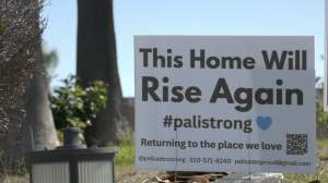 A white yard sign stands on a patch of grass in front of trees. The sign reads, “This Home Will Rise Again #palistrong” with a small blue heart. Below it says, “Returning to the place we love,” along with contact information and a QR code. A small outdoor light fixture sits in the foreground.