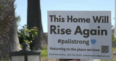 A white yard sign stands on a patch of grass in front of trees. The sign reads, “This Home Will Rise Again #palistrong” with a small blue heart. Below it says, “Returning to the place we love,” along with contact information and a QR code. A small outdoor light fixture sits in the foreground.