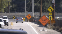 A two-lane road narrows due to construction, with multiple orange warning signs including “One Lane Road Ahead” and a flagger symbol. Cars drive through the work zone, which is bordered by barriers, fencing, and utility poles. A left-curve sign and a 40-mph speed limit sign are also visible.