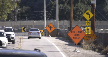 A two-lane road narrows due to construction, with multiple orange warning signs including “One Lane Road Ahead” and a flagger symbol. Cars drive through the work zone, which is bordered by barriers, fencing, and utility poles. A left-curve sign and a 40-mph speed limit sign are also visible.