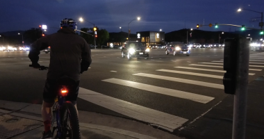 A cyclist wearing a helmet waits at a large, well-lit intersection at dusk. The wide crosswalk lines stretch across multiple lanes of traffic, where cars with headlights on move through the green light. Streetlights and buildings illuminate the scene against the darkening sky.