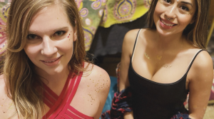 Two women smile at the camera in a warmly lit setting. The woman on the left wears a red strappy top with gold glitter on her shoulder and face. The woman on the right wears a black tank top with gold decorative designs on her face and upper chest. Colorful patterned fabric hangs in the background.