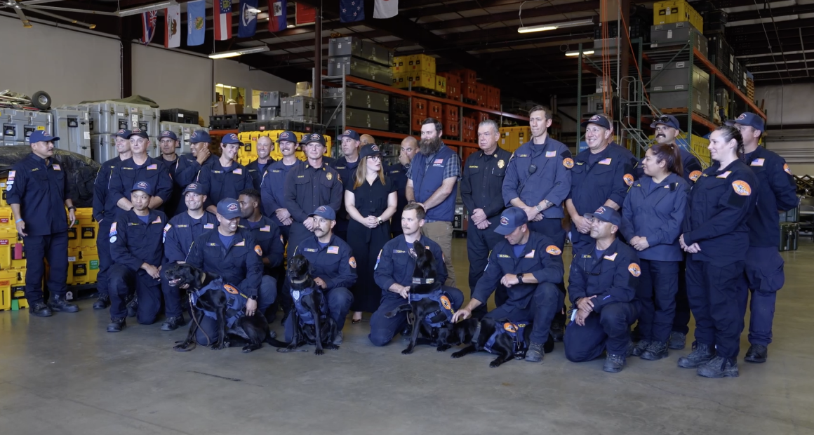 A large group of uniformed emergency response personnel pose for a group photo inside a warehouse filled with equipment cases and storage racks. Several members kneel in front holding black search-and-rescue dogs wearing harnesses. Others stand behind them, smiling, with flags hanging from the ceiling in the background.