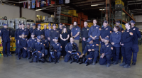 A large group of uniformed emergency response personnel pose for a group photo inside a warehouse filled with equipment cases and storage racks. Several members kneel in front holding black search-and-rescue dogs wearing harnesses. Others stand behind them, smiling, with flags hanging from the ceiling in the background.