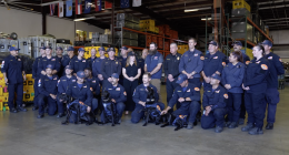 A large group of uniformed emergency response personnel pose for a group photo inside a warehouse filled with equipment cases and storage racks. Several members kneel in front holding black search-and-rescue dogs wearing harnesses. Others stand behind them, smiling, with flags hanging from the ceiling in the background.