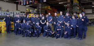 A large group of uniformed emergency response personnel pose for a group photo inside a warehouse filled with equipment cases and storage racks. Several members kneel in front holding black search-and-rescue dogs wearing harnesses. Others stand behind them, smiling, with flags hanging from the ceiling in the background.