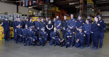 A large group of uniformed emergency response personnel pose for a group photo inside a warehouse filled with equipment cases and storage racks. Several members kneel in front holding black search-and-rescue dogs wearing harnesses. Others stand behind them, smiling, with flags hanging from the ceiling in the background.