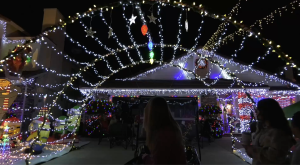 “A house decorated with an extensive display of Christmas lights, including hanging stars, colorful bulbs, and strings of white and yellow lights forming an archway over the driveway. Festive inflatables and cut-outs, such as cartoon characters in holiday outfits, sit on one side. Several people stand or sit in the foreground, looking toward the brightly lit house at night.”