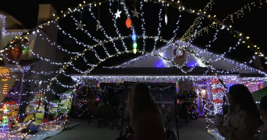 “A house decorated with an extensive display of Christmas lights, including hanging stars, colorful bulbs, and strings of white and yellow lights forming an archway over the driveway. Festive inflatables and cut-outs, such as cartoon characters in holiday outfits, sit on one side. Several people stand or sit in the foreground, looking toward the brightly lit house at night.”