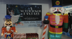 “A holiday display featuring two large nutcracker decorations—one illuminated with glittering lights on the left and a tall inflatable nutcracker on the right. Between them is a poster for ‘A Nutcracker Fantasy,’ showing a girl placing a nutcracker ornament on a Christmas tree. The poster lists showtimes for December 12 and 13 and includes a QR code for tickets. Gift-wrapped donation boxes sit below the poster, and people are visible in the background near an entrance.”