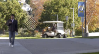 A person walks along a paved campus walkway while looking at a phone, with a maintenance golf cart driving behind them. Trees with autumn foliage, campus signage, and a blue banner line the path in the background.