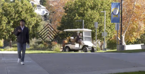 A person walks along a paved campus walkway while looking at a phone, with a maintenance golf cart driving behind them. Trees with autumn foliage, campus signage, and a blue banner line the path in the background.