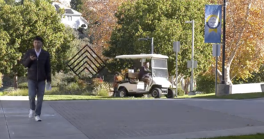 A person walks along a paved campus walkway while looking at a phone, with a maintenance golf cart driving behind them. Trees with autumn foliage, campus signage, and a blue banner line the path in the background.
