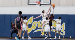 Alt text: College of the Canyons men’s basketball players in white leap to contest a shot at the rim as an opposing player in a black jersey attempts a layup, with multiple players watching beneath the basket inside a gym.