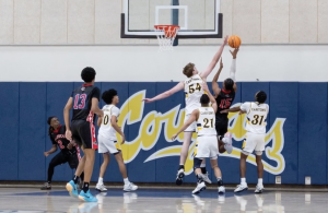 Alt text: College of the Canyons men’s basketball players in white leap to contest a shot at the rim as an opposing player in a black jersey attempts a layup, with multiple players watching beneath the basket inside a gym.
