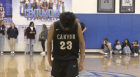 A young male basketball player wearing a black and green jersey with the word CANYON and the number 23 on the back stands in the center of a gymnasium court. He is looking down and appears to be concentrating or dejected. In the background, there are spectators sitting on the court and standing along the edges. A large blue banner or sign behind him reads CENTURIONS in white text, and there are framed jerseys or memorabilia on the blue wall to the right. The floor is a wooden basketball court with markings visible, including a large graphic under the player's feet.