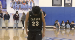 A young male basketball player wearing a black and green jersey with the word CANYON and the number 23 on the back stands in the center of a gymnasium court. He is looking down and appears to be concentrating or dejected. In the background, there are spectators sitting on the court and standing along the edges. A large blue banner or sign behind him reads CENTURIONS in white text, and there are framed jerseys or memorabilia on the blue wall to the right. The floor is a wooden basketball court with markings visible, including a large graphic under the player's feet.