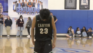 A young male basketball player wearing a black and green jersey with the word CANYON and the number 23 on the back stands in the center of a gymnasium court. He is looking down and appears to be concentrating or dejected. In the background, there are spectators sitting on the court and standing along the edges. A large blue banner or sign behind him reads CENTURIONS in white text, and there are framed jerseys or memorabilia on the blue wall to the right. The floor is a wooden basketball court with markings visible, including a large graphic under the player's feet.