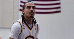 Donovan Chapman in a white jersey with a large “C” on the front focuses intently while preparing to shoot, with the American flag hanging on the wall behind him.