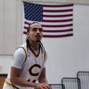 Donovan Chapman in a white jersey with a large “C” on the front focuses intently while preparing to shoot, with the American flag hanging on the wall behind him.