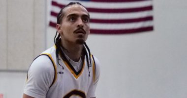 Donovan Chapman in a white jersey with a large “C” on the front focuses intently while preparing to shoot, with the American flag hanging on the wall behind him.