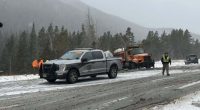 A snowy, slush-covered stretch of Interstate 70 in a mountain area, with a state patrol pickup truck parked in the roadway, an orange snowplow stopped behind it, and emergency responders standing nearby as light snow falls and evergreen-covered hills rise in the background