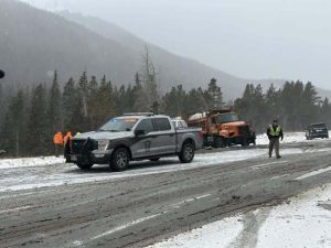 A snowy, slush-covered stretch of Interstate 70 in a mountain area, with a state patrol pickup truck parked in the roadway, an orange snowplow stopped behind it, and emergency responders standing nearby as light snow falls and evergreen-covered hills rise in the background