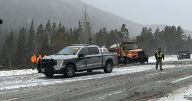 A snowy, slush-covered stretch of Interstate 70 in a mountain area, with a state patrol pickup truck parked in the roadway, an orange snowplow stopped behind it, and emergency responders standing nearby as light snow falls and evergreen-covered hills rise in the background
