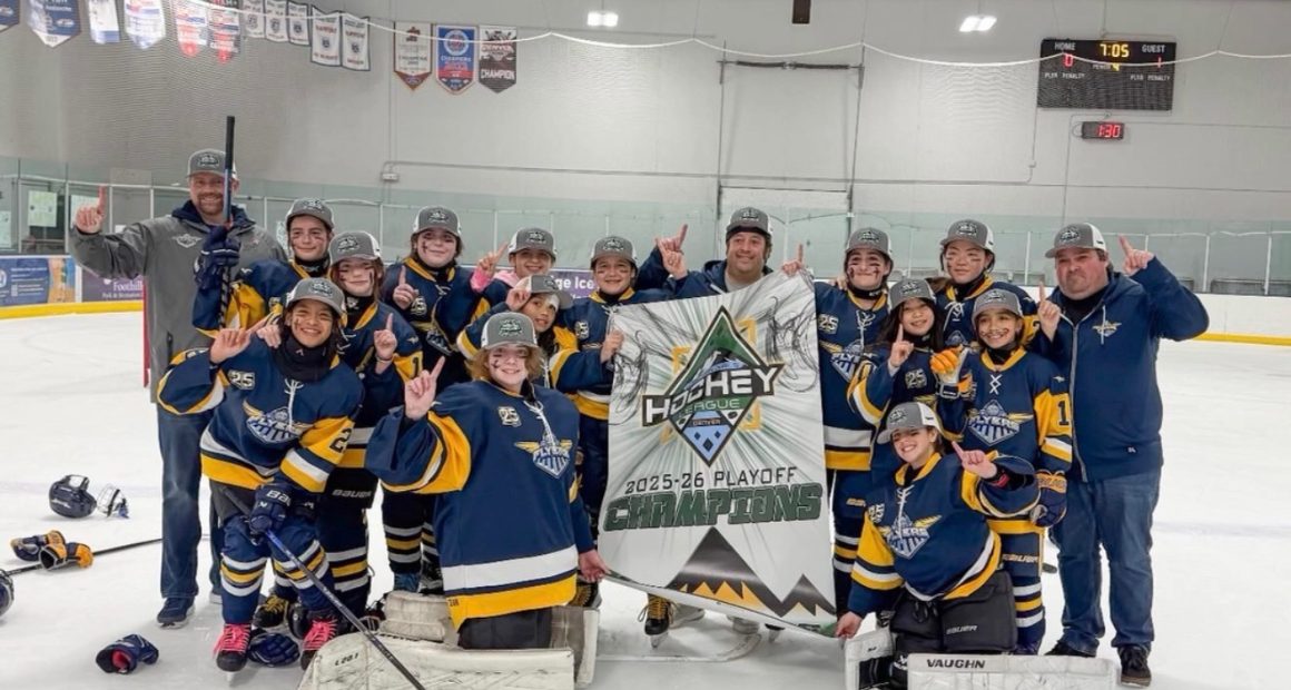 **Alt text:** A youth girls hockey team in blue and gold uniforms poses on the ice with coaches, smiling and raising one finger while holding a banner that reads “Hockey House 2025–26 Playoff Champions” inside an indoor rink.