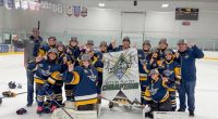**Alt text:** A youth girls hockey team in blue and gold uniforms poses on the ice with coaches, smiling and raising one finger while holding a banner that reads “Hockey House 2025–26 Playoff Champions” inside an indoor rink.