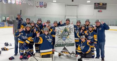 **Alt text:** A youth girls hockey team in blue and gold uniforms poses on the ice with coaches, smiling and raising one finger while holding a banner that reads “Hockey House 2025–26 Playoff Champions” inside an indoor rink.