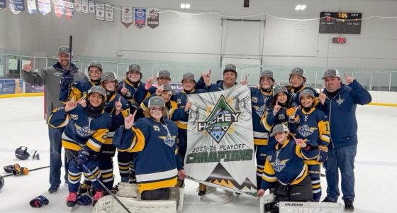 **Alt text:** A youth girls hockey team in blue and gold uniforms poses on the ice with coaches, smiling and raising one finger while holding a banner that reads “Hockey House 2025–26 Playoff Champions” inside an indoor rink.