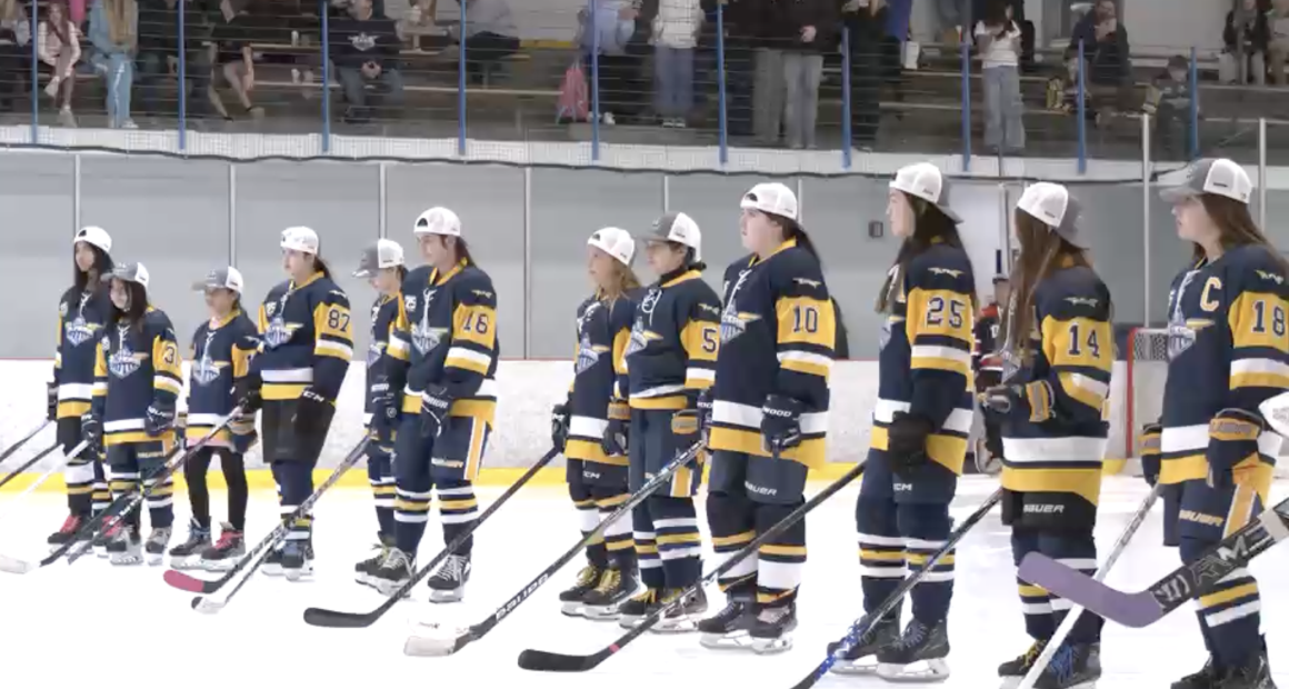 A youth girls’ hockey team stands shoulder to shoulder on the ice wearing navy and gold jerseys and white championship caps, holding their sticks during a postgame ceremony as spectators watch from the stands behind them.