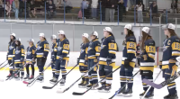 A youth girls’ hockey team stands shoulder to shoulder on the ice wearing navy and gold jerseys and white championship caps, holding their sticks during a postgame ceremony as spectators watch from the stands behind them.