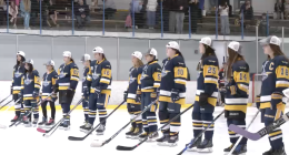 A youth girls’ hockey team stands shoulder to shoulder on the ice wearing navy and gold jerseys and white championship caps, holding their sticks during a postgame ceremony as spectators watch from the stands behind them.