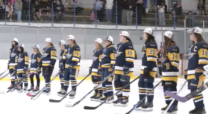 A youth girls’ hockey team stands shoulder to shoulder on the ice wearing navy and gold jerseys and white championship caps, holding their sticks during a postgame ceremony as spectators watch from the stands behind them.