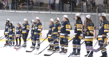 A youth girls’ hockey team stands shoulder to shoulder on the ice wearing navy and gold jerseys and white championship caps, holding their sticks during a postgame ceremony as spectators watch from the stands behind them.