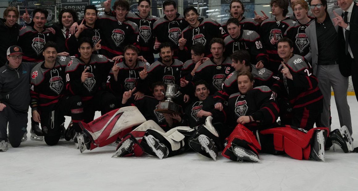 University of New Mexico Lobos in black and red uniforms poses on the ice with a championship trophy, celebrating at The Cube rink after a playoff win