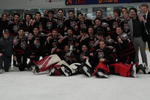 University of New Mexico Lobos in black and red uniforms poses on the ice with a championship trophy, celebrating at The Cube rink after a playoff win